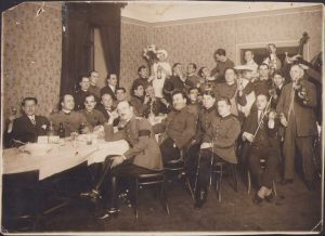 Viitorul general Nicolae Macici (cu monoclu negru), în poză cu gradul de căpitan, purtând două însemne de rănit pe mânecă, alături de alți ofițeri la petrecere cu lăutari